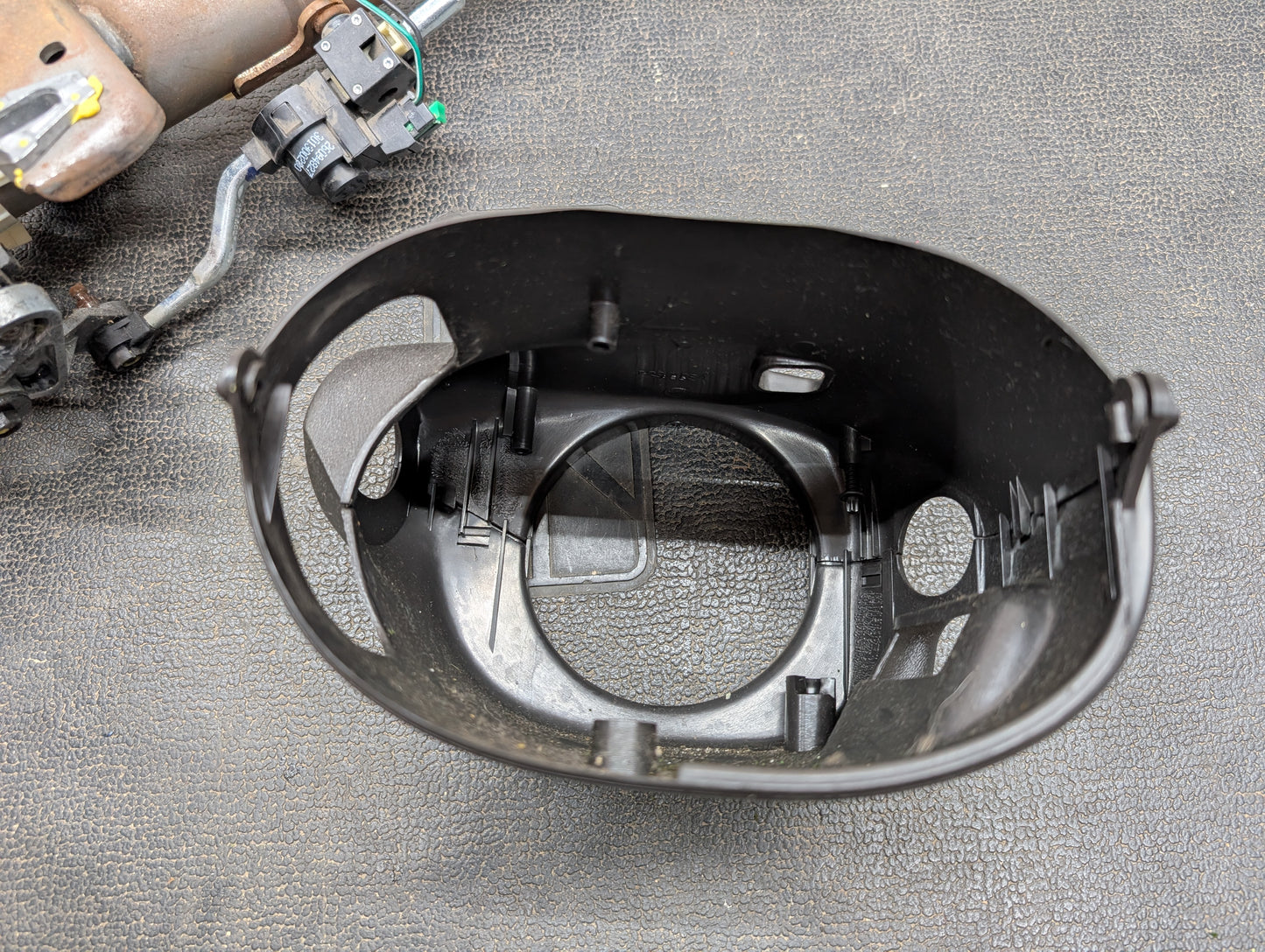 A close-up of a black plastic automotive part on a dark textured surface, with metal and plastic components including a General Motors OEM Steering Column w/Tilt, Cruise Control, and Column Shift for 1998-2005 Chevy S10 Blazer visible to the left.