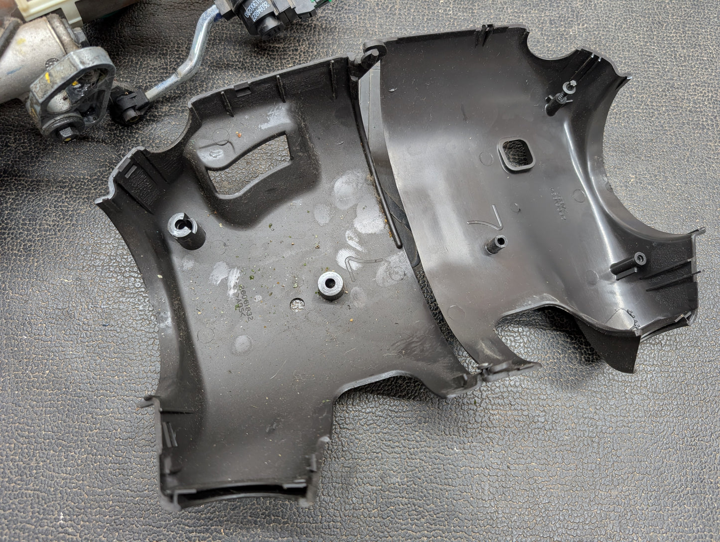An open black plastic cover rests on a textured surface, revealing part of an OEM Steering Column with Tilt, Cruise Control, and Column Shift for 1998-2005 Chevy S10 Blazer by General Motors in the upper left corner.