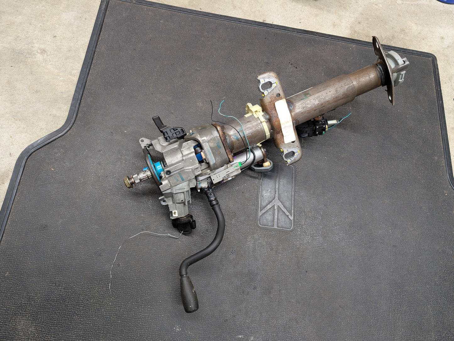 A General Motors OEM Steering Column with tilt, cruise control, and column shift for 1998-2005 Chevy S10, Blazer, and more is shown resting on a black textured mat placed on a concrete floor.