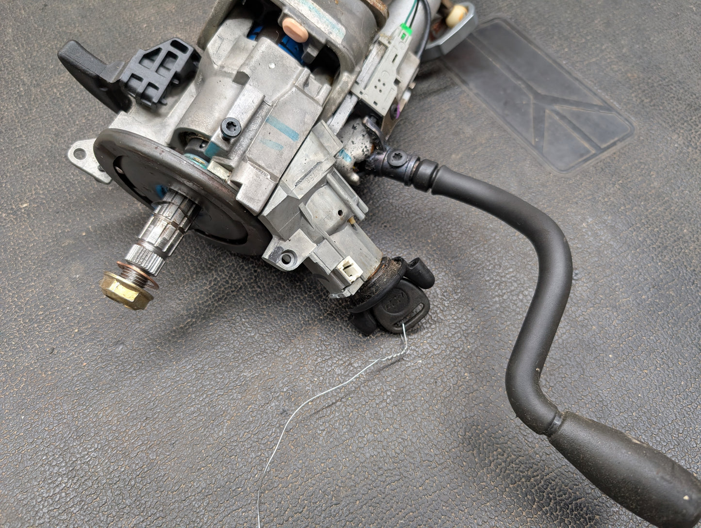 A General Motors OEM Steering Column with tilt, cruise control, and column shift for 1998-2005 Chevy S10 Blazer models is shown close-up with key sensor and visible mechanical/electronic parts on a textured surface.
