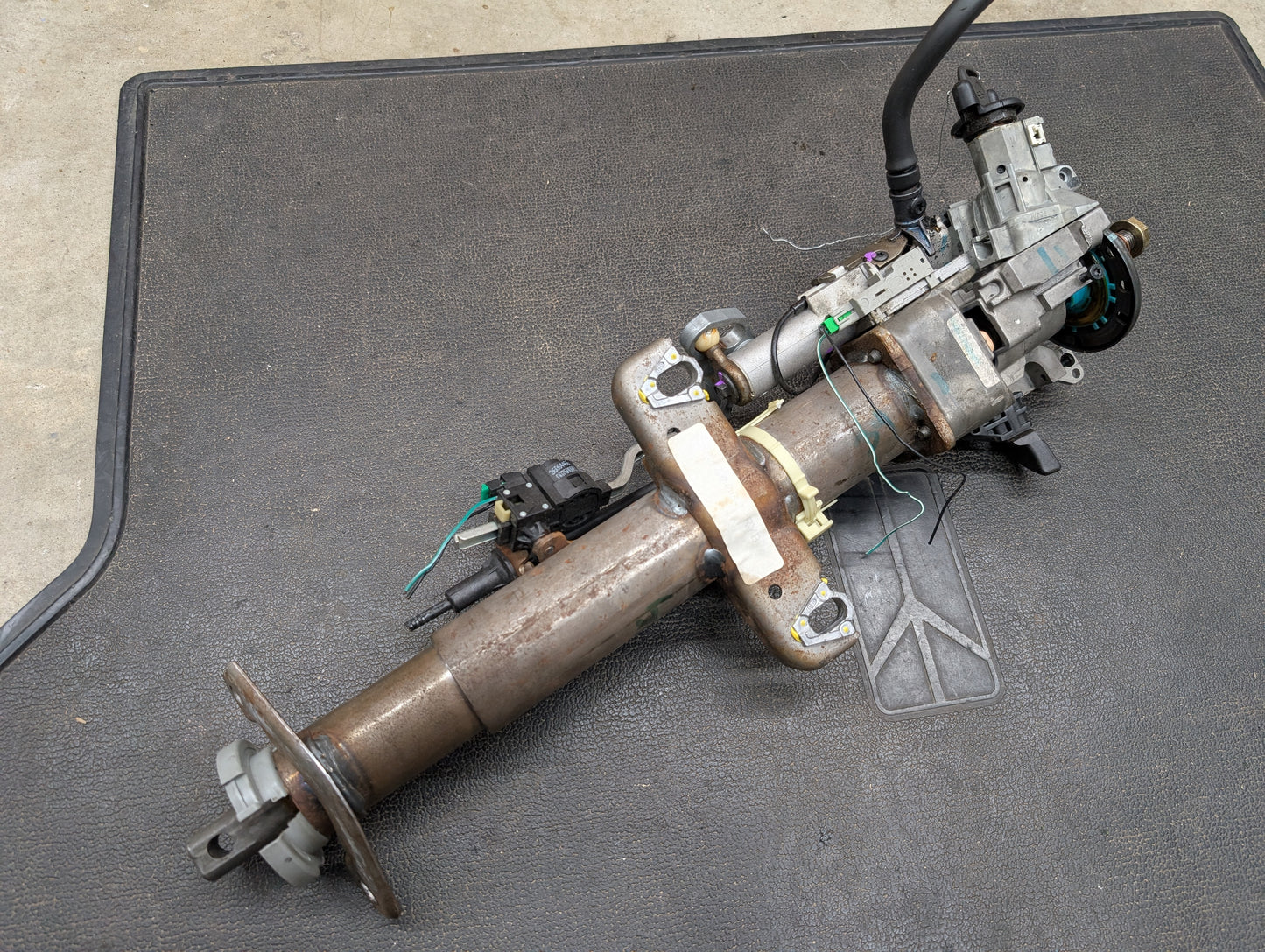 A General Motors OEM Steering Column with tilt, cruise control, and column shift for 1998-2005 Chevy S10/Blazer is displayed on a textured black mat, viewed from above.