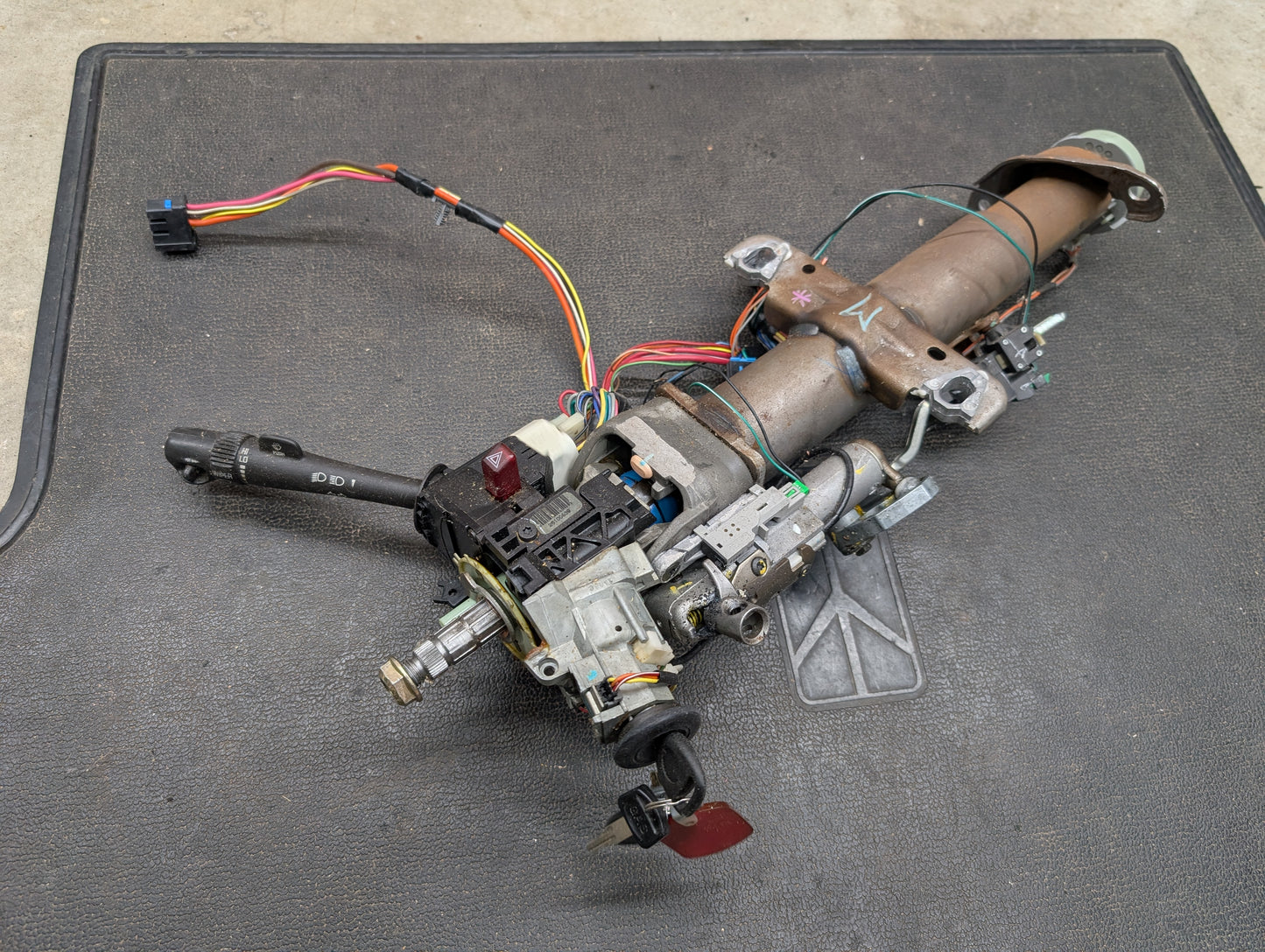 A General Motors OEM Steering Column w/KEY for 2000-2002 GM Full Size Trucks is shown with wiring, ignition switch, and turn signal lever attached, designed for automatic transmissions, on a black textured mat.
