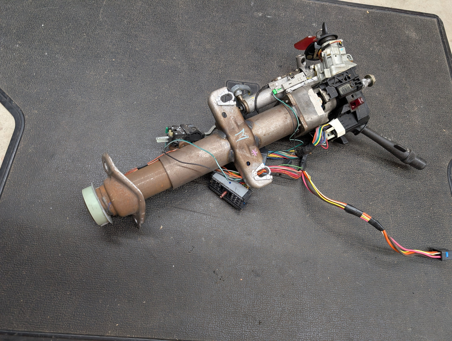 A General Motors OEM Steering Column w/KEY for 2000-2002 GM Full Size Trucks, featuring attached wiring, levers, and switches for automatic transmission, rests on a black textured surface. The assembly includes metal and plastic components.