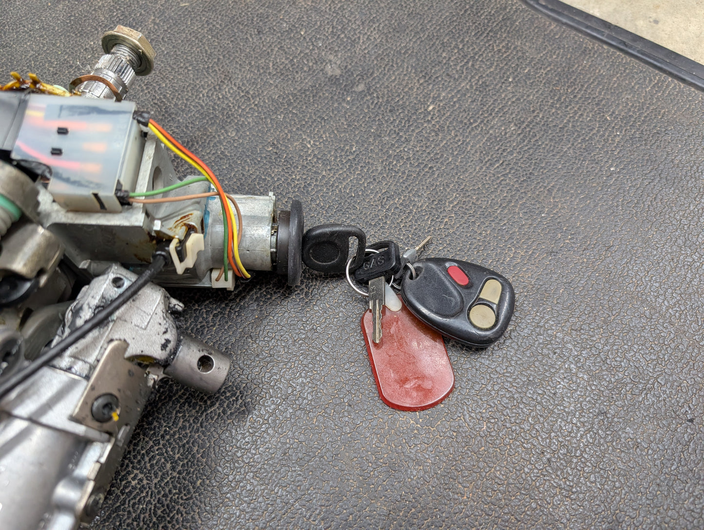 A General Motors OEM Steering Column w/KEY for 2000-2002 GM Full Size Trucks is shown with car keys, a black remote, metal keys, and a red tag on a textured black surface; wiring is attached to the ignition switch mechanism.