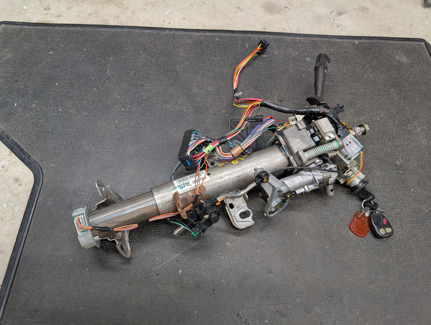 A General Motors OEM Steering Column w/KEY for 2000-2002 GM Full Size Trucks, including wiring and ignition switch for automatic transmission, is shown on a black mat in a workshop setting.
