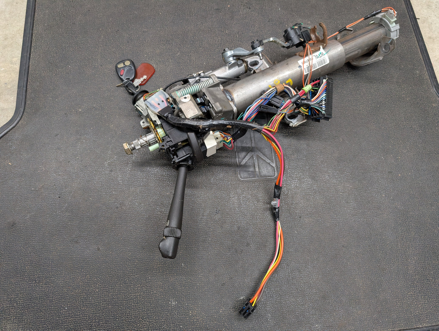 A General Motors OEM Steering Column w/KEY for 2000-2002 GM Full Size Trucks, including ignition switch, wiring harnesses, and connectors, is shown on a black textured mat.