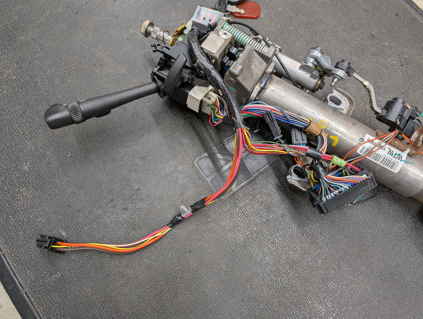A close-up of a General Motors OEM Steering Column w/KEY for 2000-2002 GM Full Size Trucks, showing colorful wires, connectors, and automatic transmission components on a black textured surface.