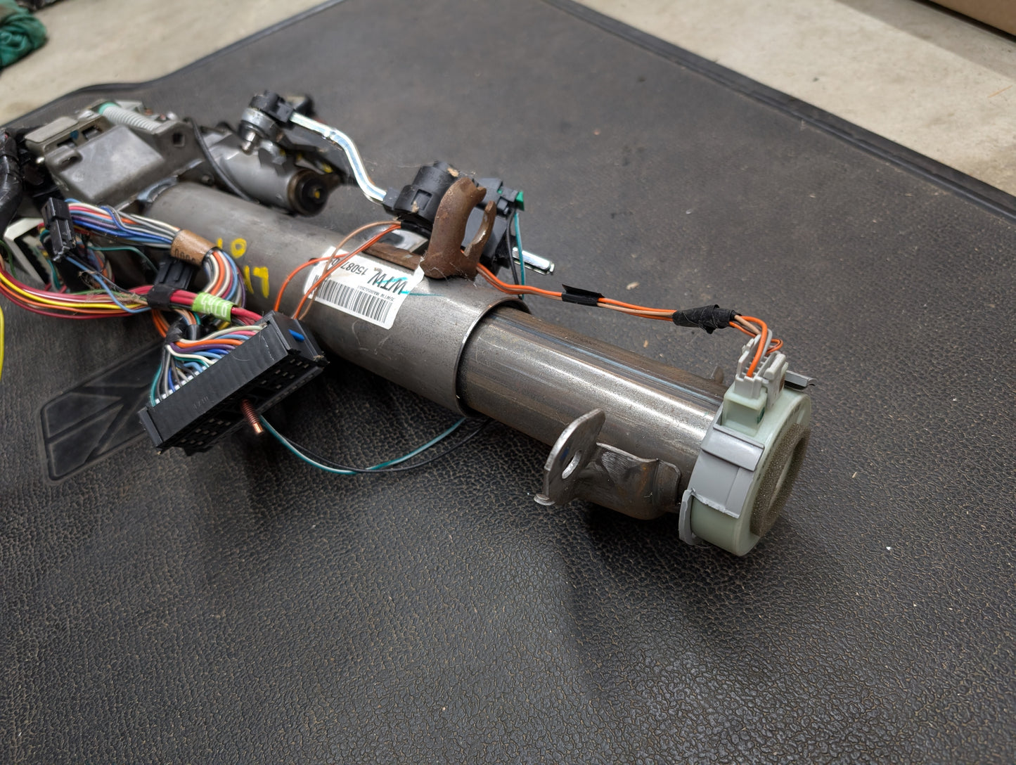 An OEM Steering Column w/KEY for 2000-2002 GM Full Size Trucks by General Motors is shown resting on a textured black surface, complete with wiring, connectors, colorful wires, and a barcode label visible on the metal tube.