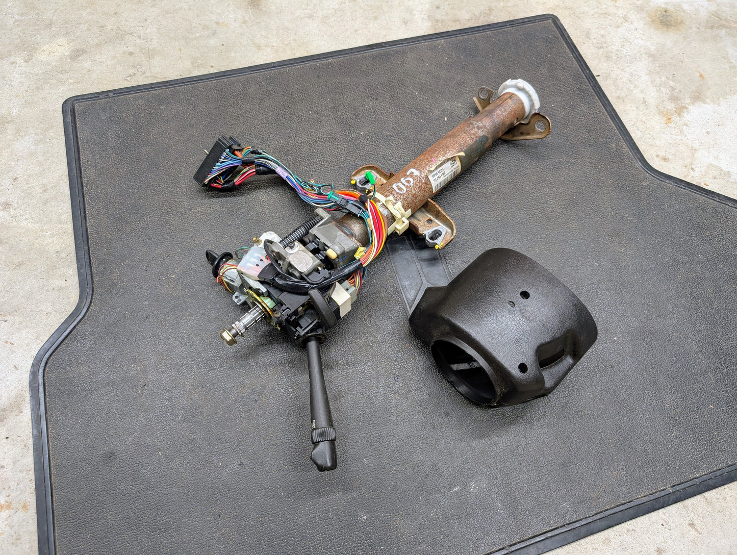 A General Motors OEM Steering Column with tilt, cruise control, and console shift for 1998-2005 Chevy S10 Blazer and more is shown on a black mat, with wiring, controls attached, and a black plastic cover beside it.