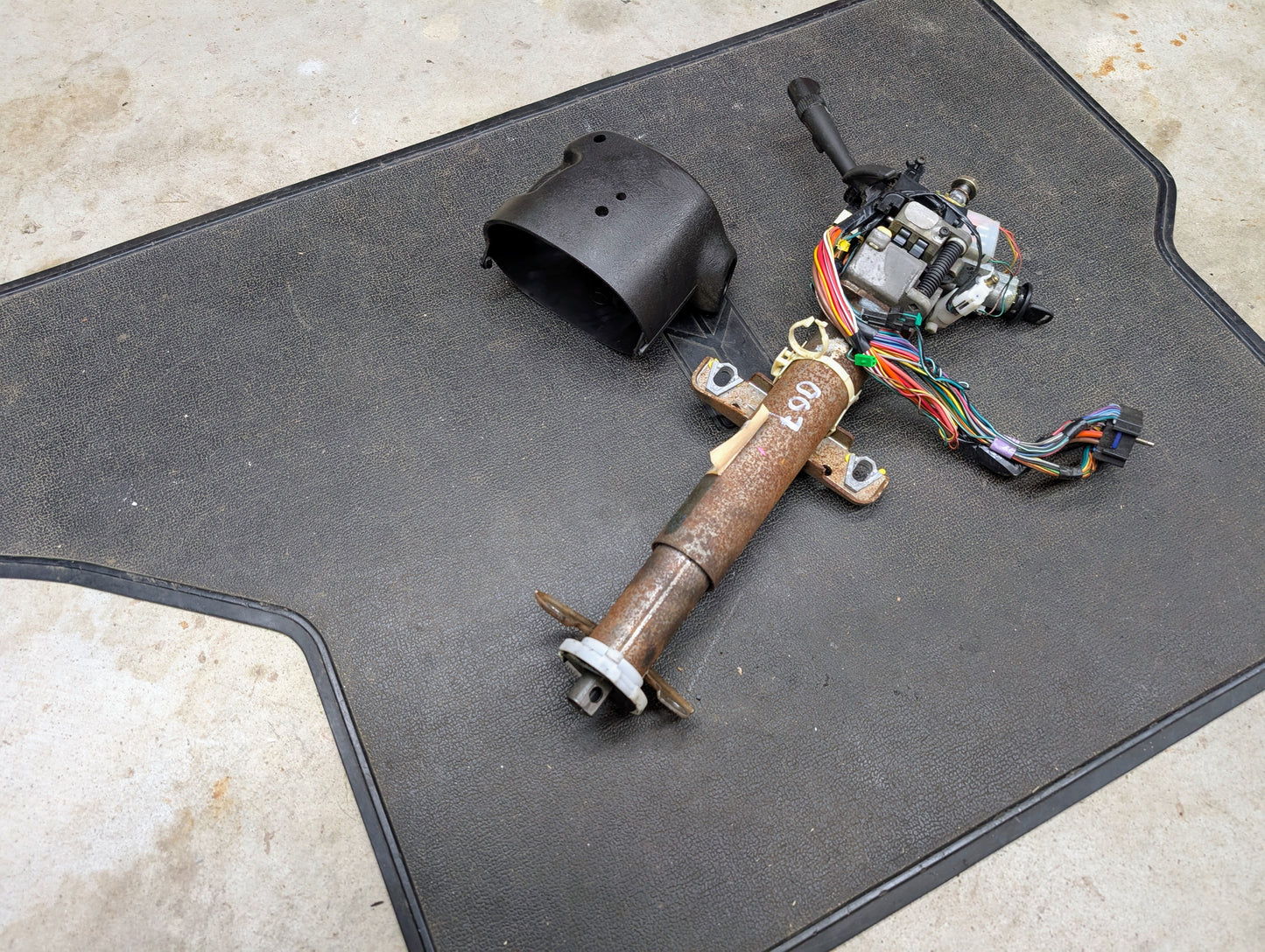 A General Motors OEM Steering Column with tilt, cruise control, and console shift for 1998-2005 Chevy S10 Blazer rests on a textured black mat atop a concrete floor, wiring and black plastic cover attached.