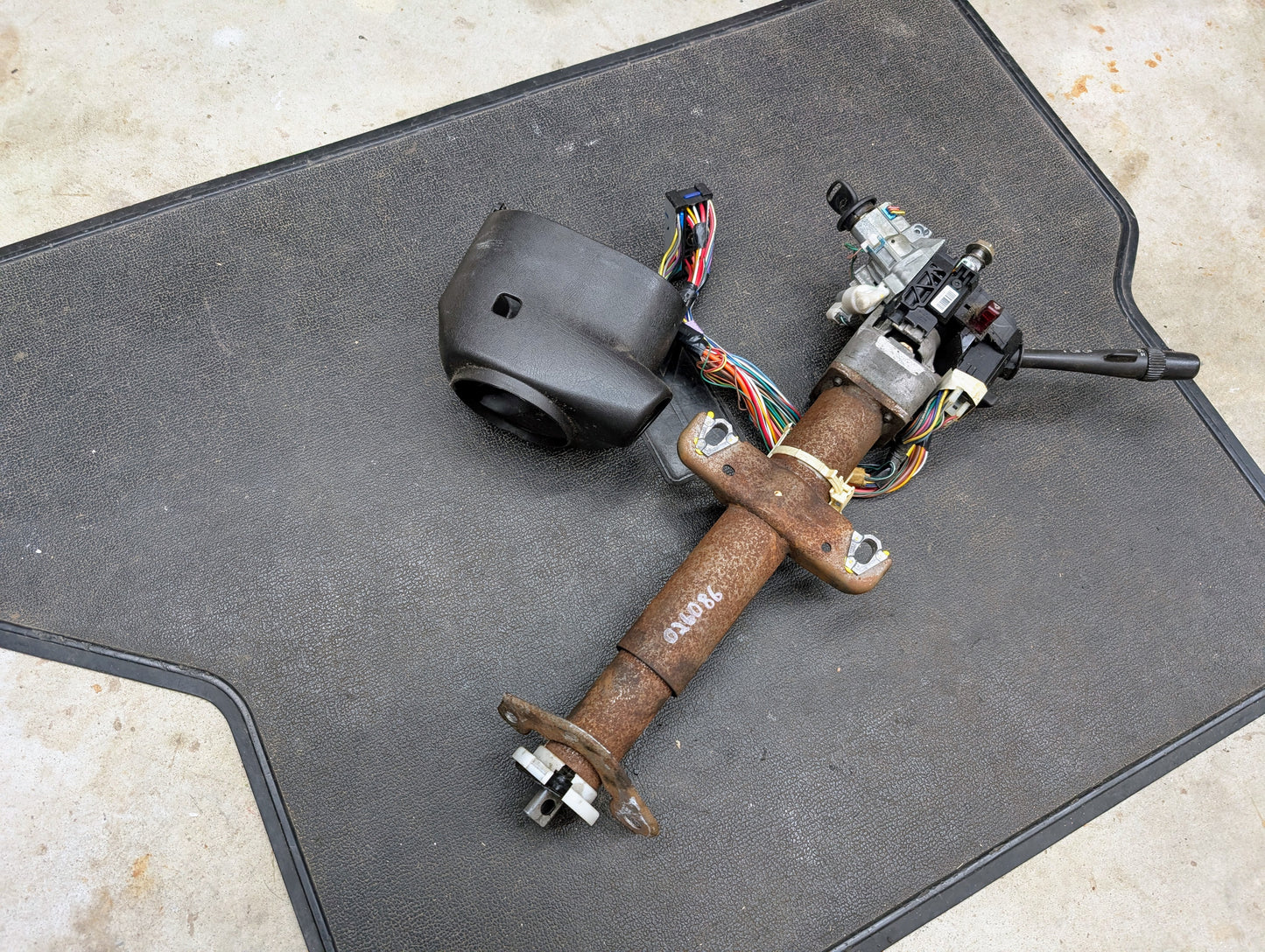 A General Motors OEM Steering Column with tilt, cruise control, and console shift for 1998-2005 Chevy S10 Blazer rests on a black floor mat on concrete, complete with wiring, ignition switch, and steering lock.