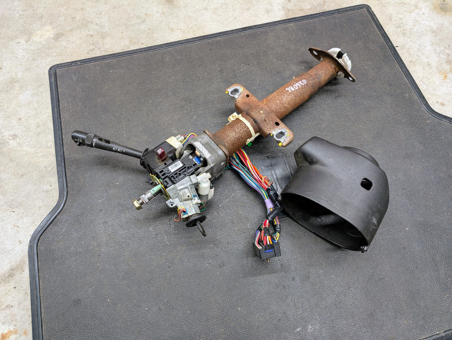 A General Motors OEM Steering Column w/Tilt, Cruise Control, and Console Shift for 1998-2005 Chevy S10 Blazer and more is shown with wiring, switches, and a separate black plastic cover on a black mat over concrete.