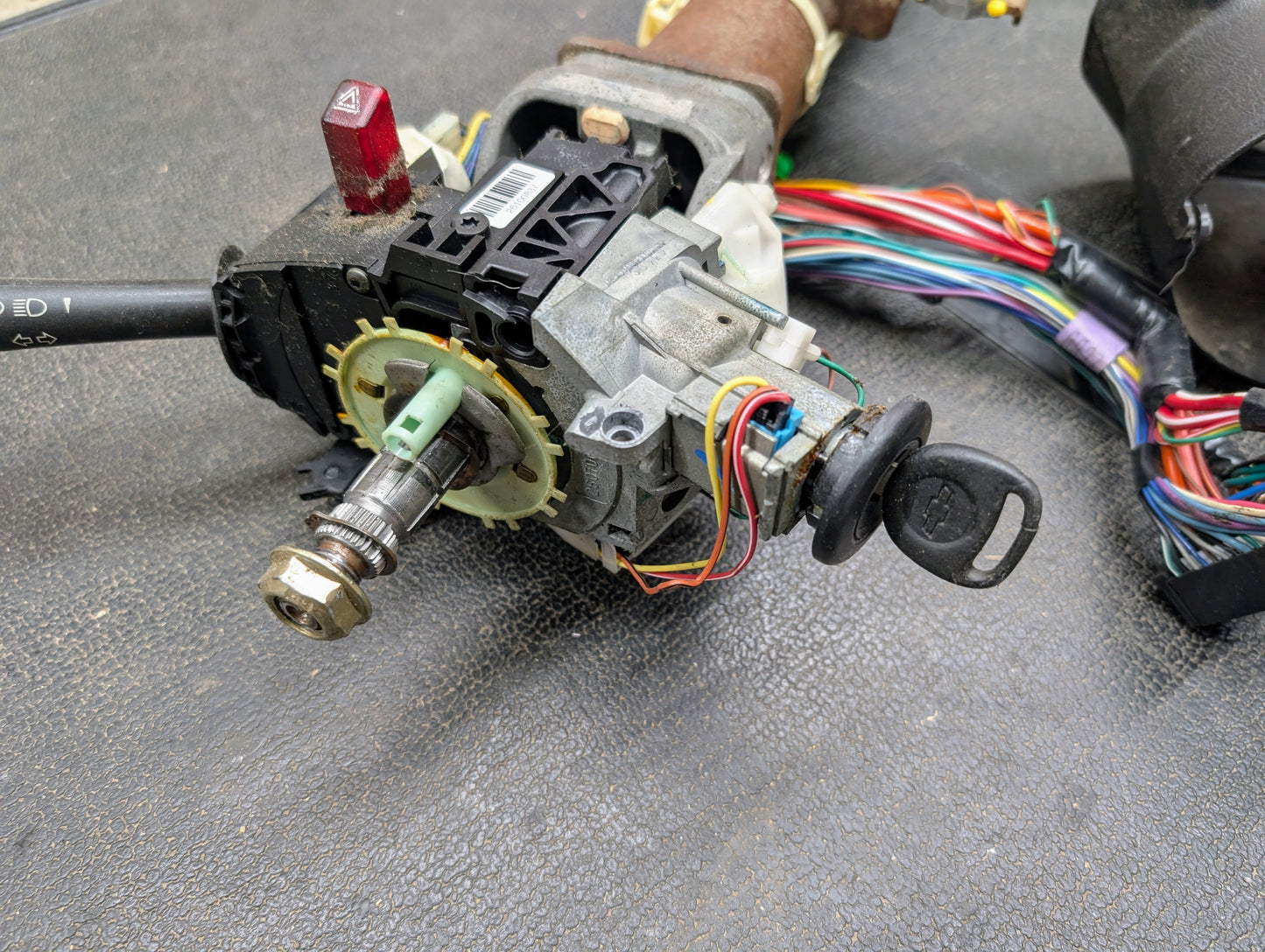 A close-up of a General Motors OEM Steering Column with tilt, cruise control, and console shift for 1998-2005 Chevy S10 Blazer and more, showing the ignition switch assembly with key, wiring harness, and mechanical components exposed.