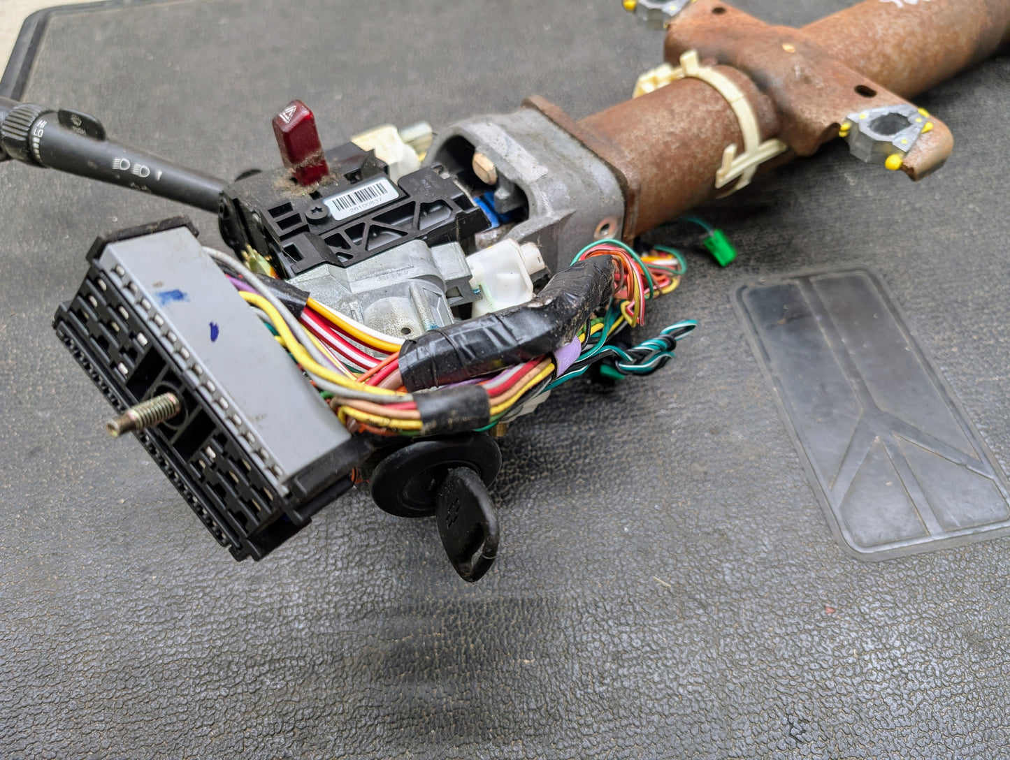 A close-up of a General Motors OEM Steering Column w/Tilt, Cruise Control, and Console Shift for 1998-2005 Chevy S10 Blazer shows exposed wiring, ignition key, switches, and black connector on a textured surface.