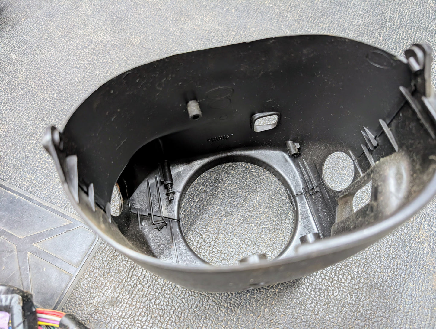 A close-up of a black plastic automotive headlight housing with mounting points rests on a textured gray surface. Wires peek out in the lower left—a perfect OEM Steering Column w/Tilt, Cruise Control, and Console Shift by General Motors for 1998-2005 Chevy S10 Blazer.