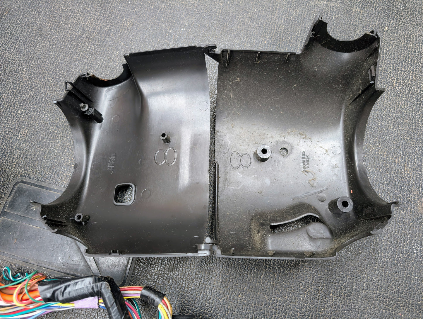 Two black plastic covers from a General Motors OEM Steering Column w/Tilt, Cruise Control, and Console Shift for 1998-2005 Chevy S10 Blazer are side by side on a textured surface, with wires and part of a control module at lower left.