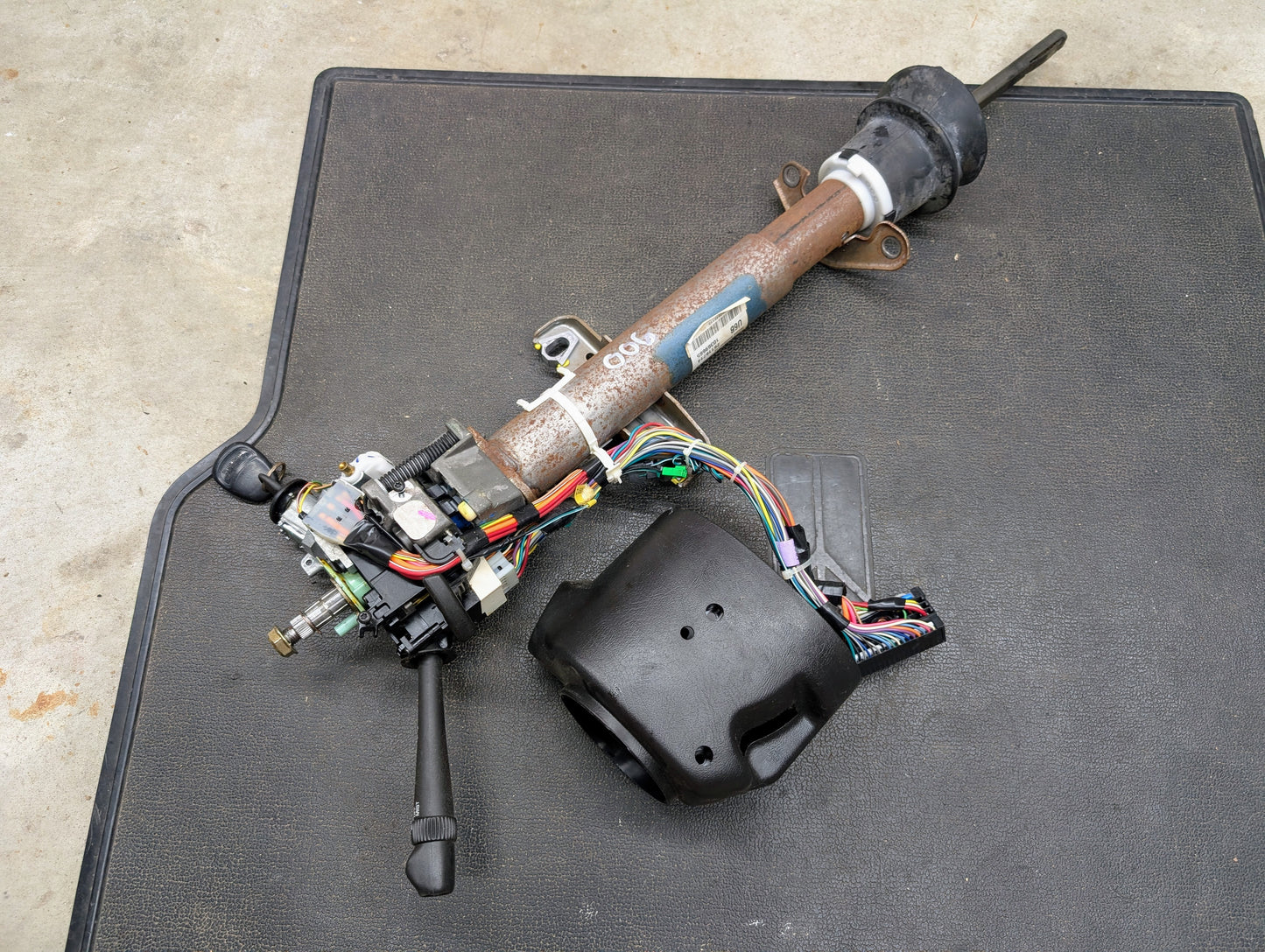 A General Motors OEM Steering Column w/Tilt, Cruise Control, and Console Shift for 1998-2005 Chevy S10 Blazer and more is shown disassembled with wiring and housing exposed on a black mat over a concrete floor.