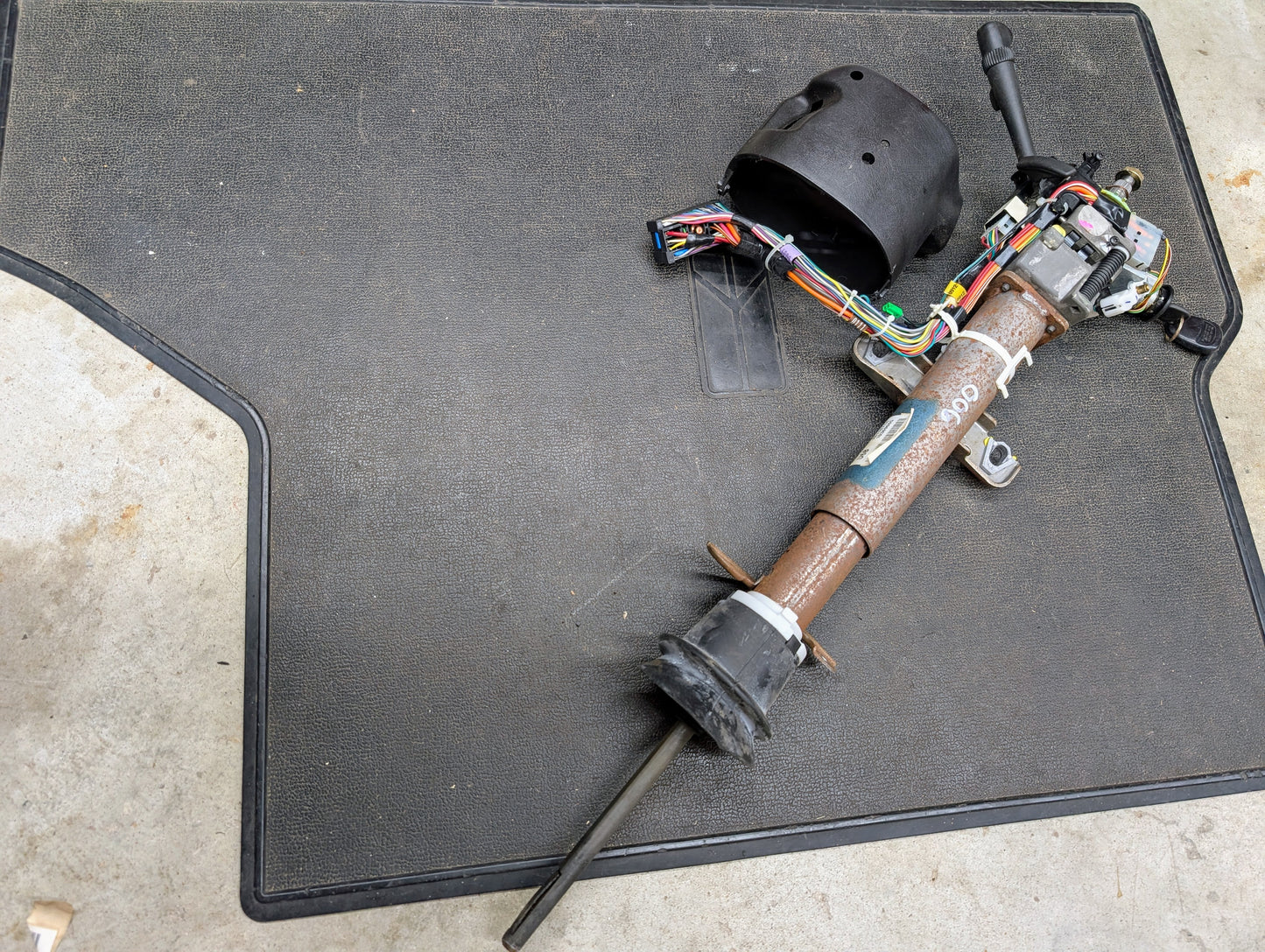 A General Motors OEM Steering Column w/Tilt, Cruise Control, and Console Shift for 1998-2005 Chevy S10 Blazer and more is disassembled with visible wiring and plastic parts on a black floor mat over concrete.