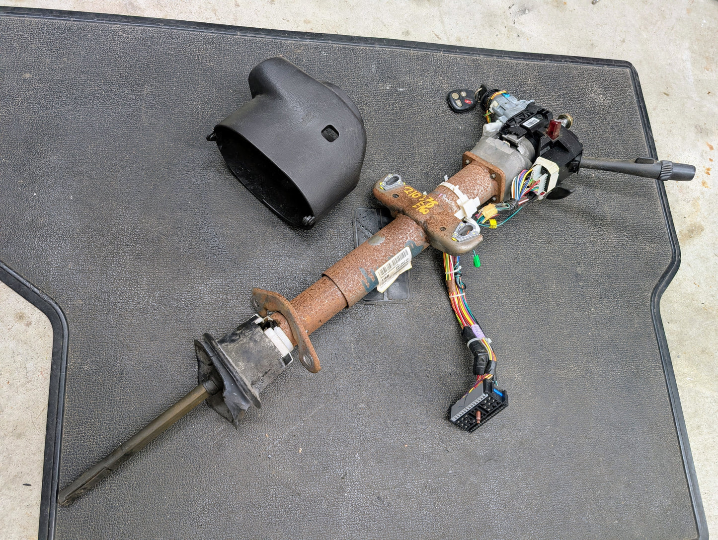 A used General Motors OEM steering column with tilt, cruise control, and console shift for 1998-2005 Chevy S10 Blazer rests on a black mat with wiring; visible rust is present and the removed black plastic cover lies beside it.