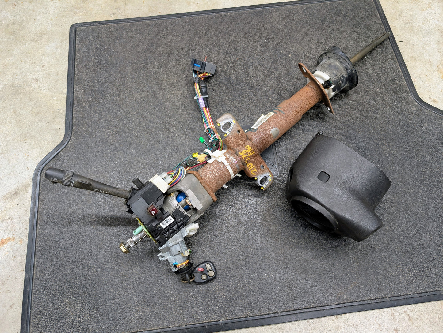 A General Motors OEM Steering Column with tilt, cruise control, and console shift for 1998-2005 Chevy S10 Blazer is shown with wiring and switches on a black mat. The black plastic column cover is also included.