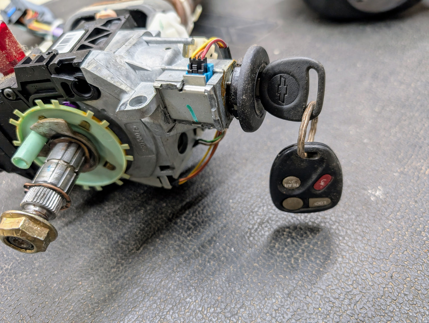 A close-up of a General Motors OEM Steering Column with tilt, cruise control, and console shift for 1998-2005 Chevy S10 Blazer, showing the ignition switch assembly with car key featuring the Chevy logo beside the column.