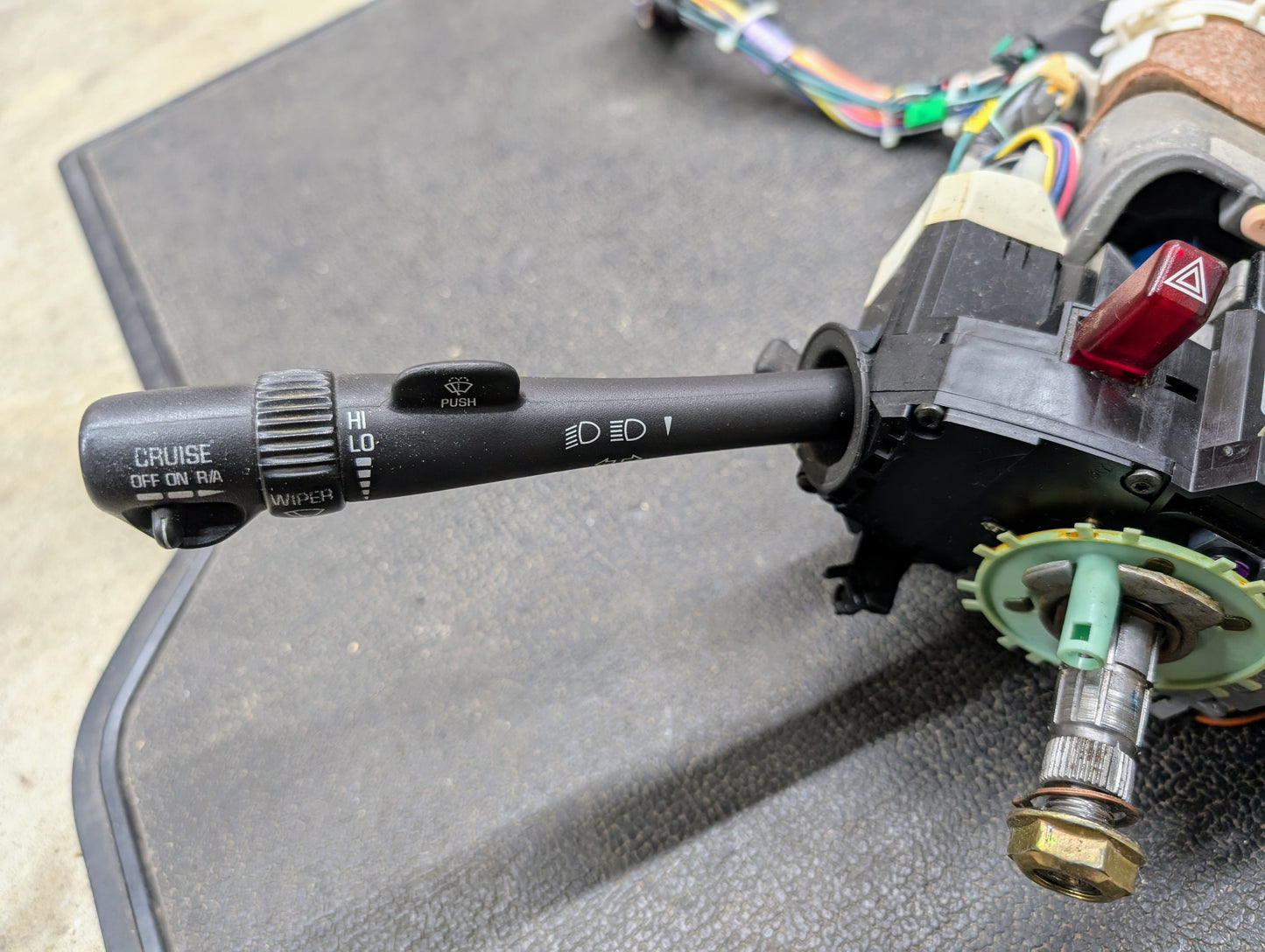 A close-up of a General Motors OEM Steering Column w/Tilt, Cruise Control, and Console Shift for 1998-2005 Chevy S10 Blazer, showing the turn signal and wiper control lever with wiring and mechanical parts on a black textured surface.