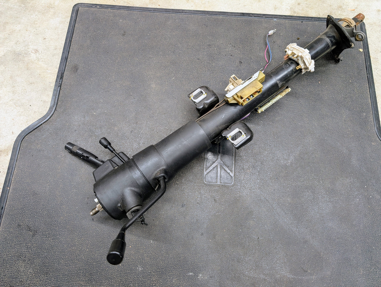 A General Motors OEM Steering Column with tilt and cruise control for 1988-1993 Chevy S10, Blazer, and more is shown on a textured mat, complete with attached wiring and switch modules.