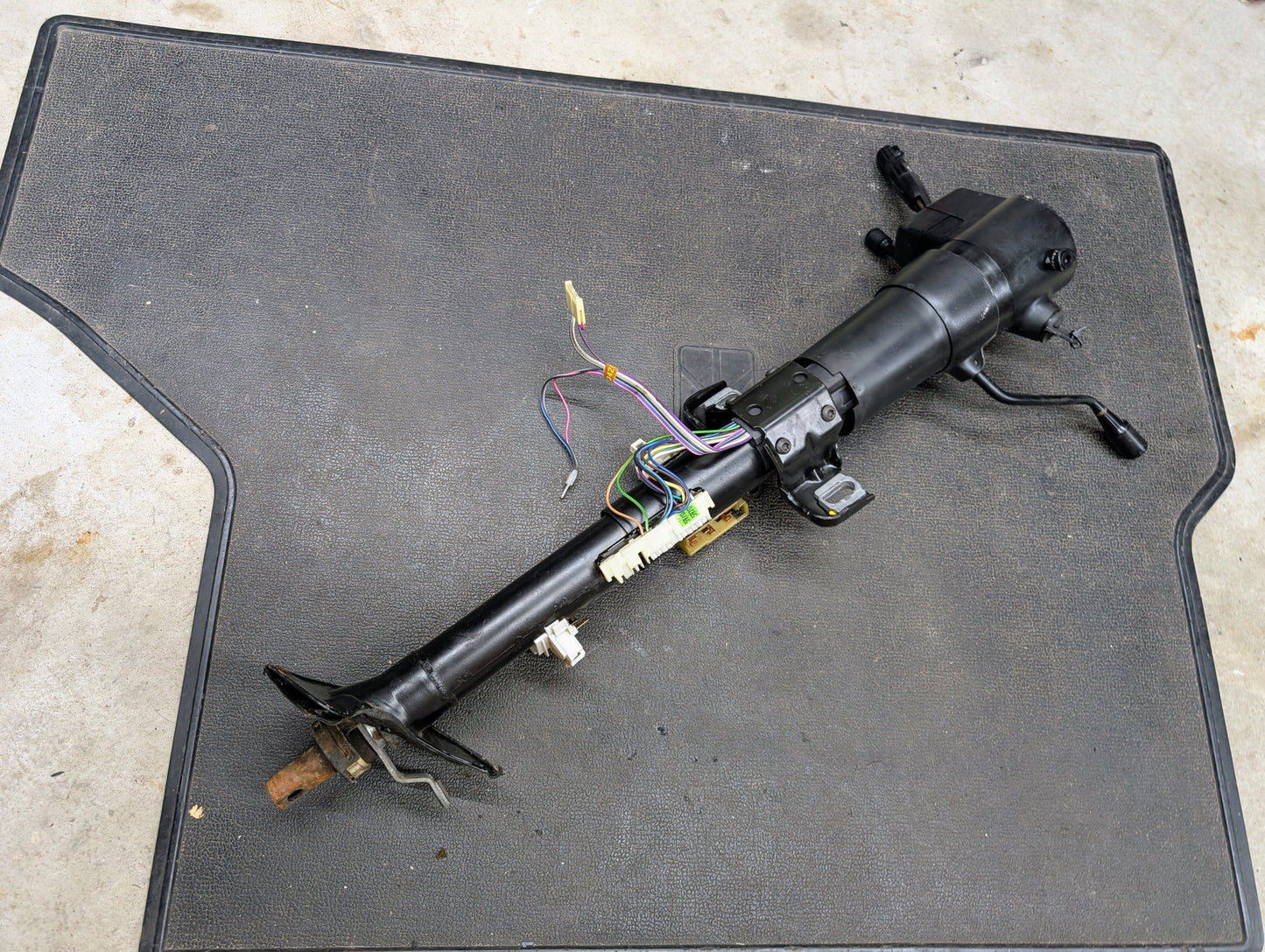A General Motors OEM Steering Column with tilt and cruise control for 1988-1993 Chevy S10, Blazer, and more is shown with exposed wires and connectors on a textured black mat atop a concrete floor.