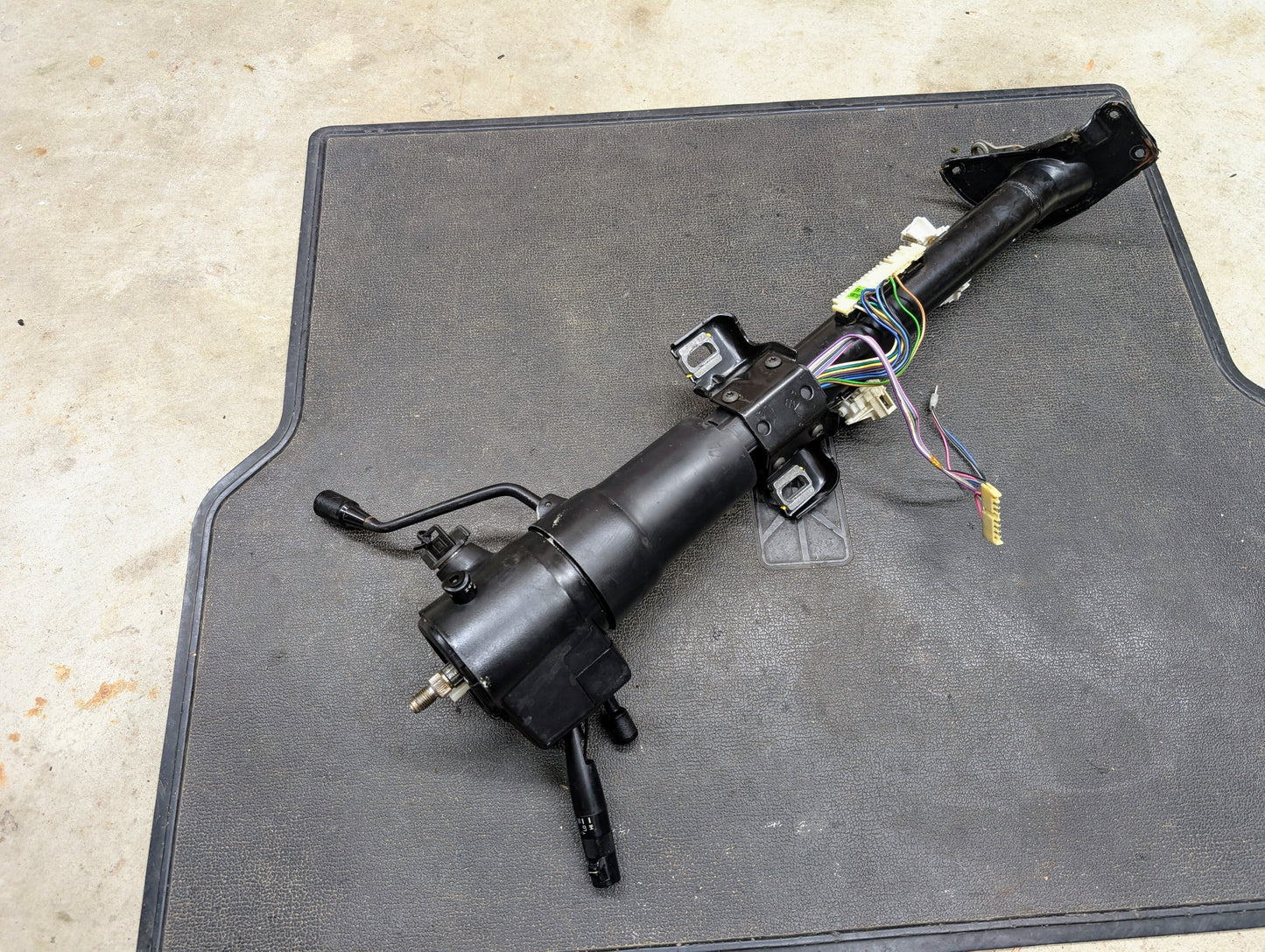 A black General Motors OEM Steering Column with tilt and cruise control for 1988-1993 Chevy S10, Blazer, and more, designed for automatic transmission models, rests on a gray mat over a concrete floor.
