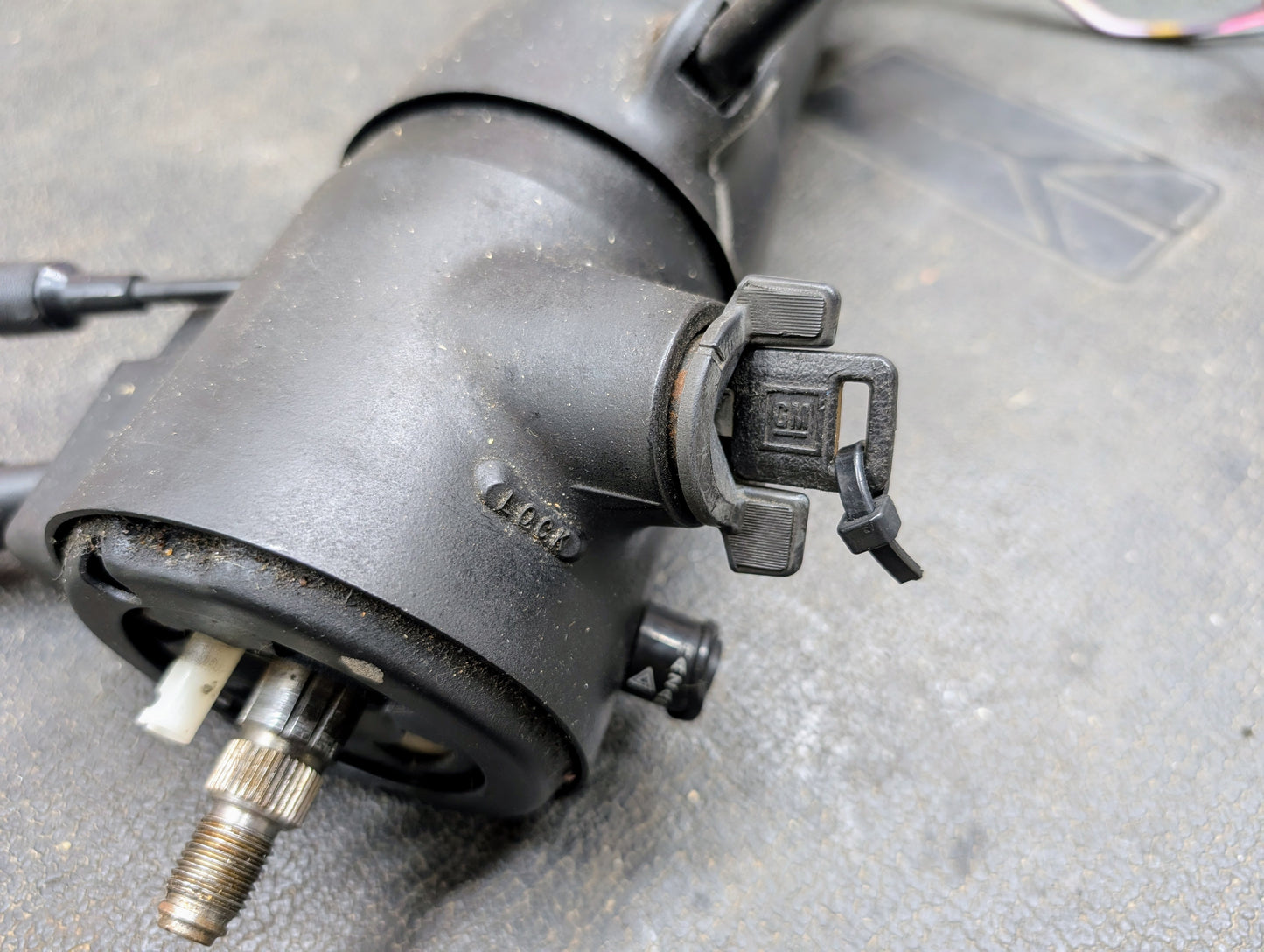 Close-up of a black mechanical component from a General Motors OEM Steering Column w/Tilt and Cruise Control for 1988-1993 Chevy S10/Blazer, showing gear shaft, molded markings, and a zip-tied plastic connector on a textured surface.