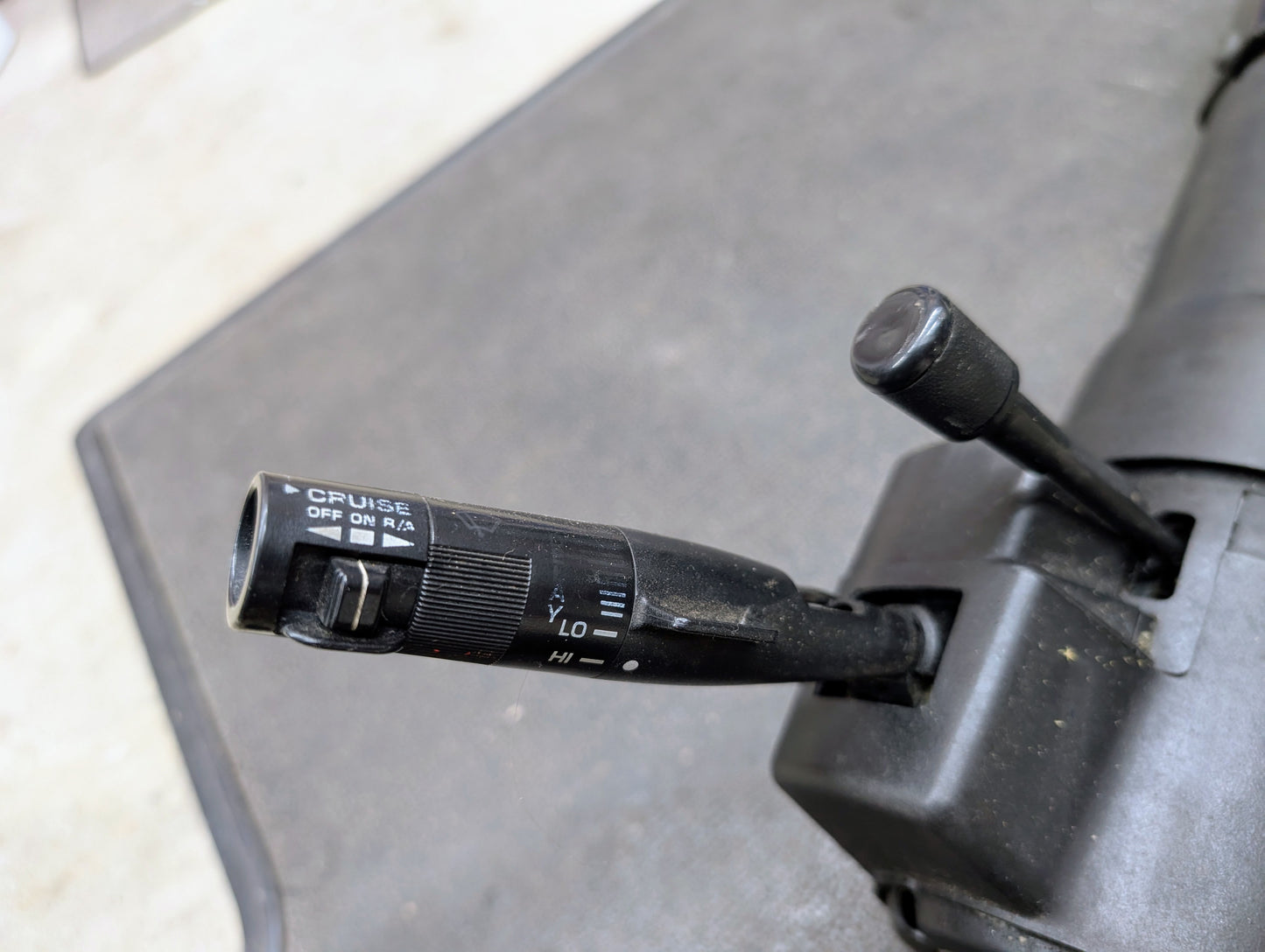Close-up of a General Motors OEM Steering Column w/Tilt and Cruise Control for 1988-1993 Chevy S10, Blazer, and more, showing cruise control and turn signal levers for automatic transmission on a black textured surface.
