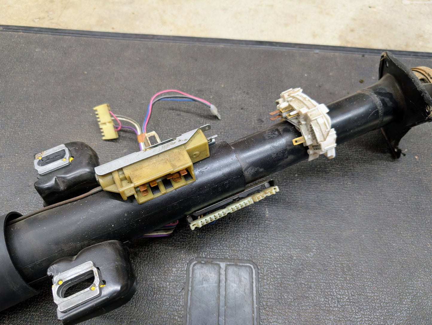 A close-up of a General Motors OEM Steering Column with tilt and cruise control for 1988-1993 Chevy S10, Blazer, and more, showing exposed wiring and connectors on a textured black surface.