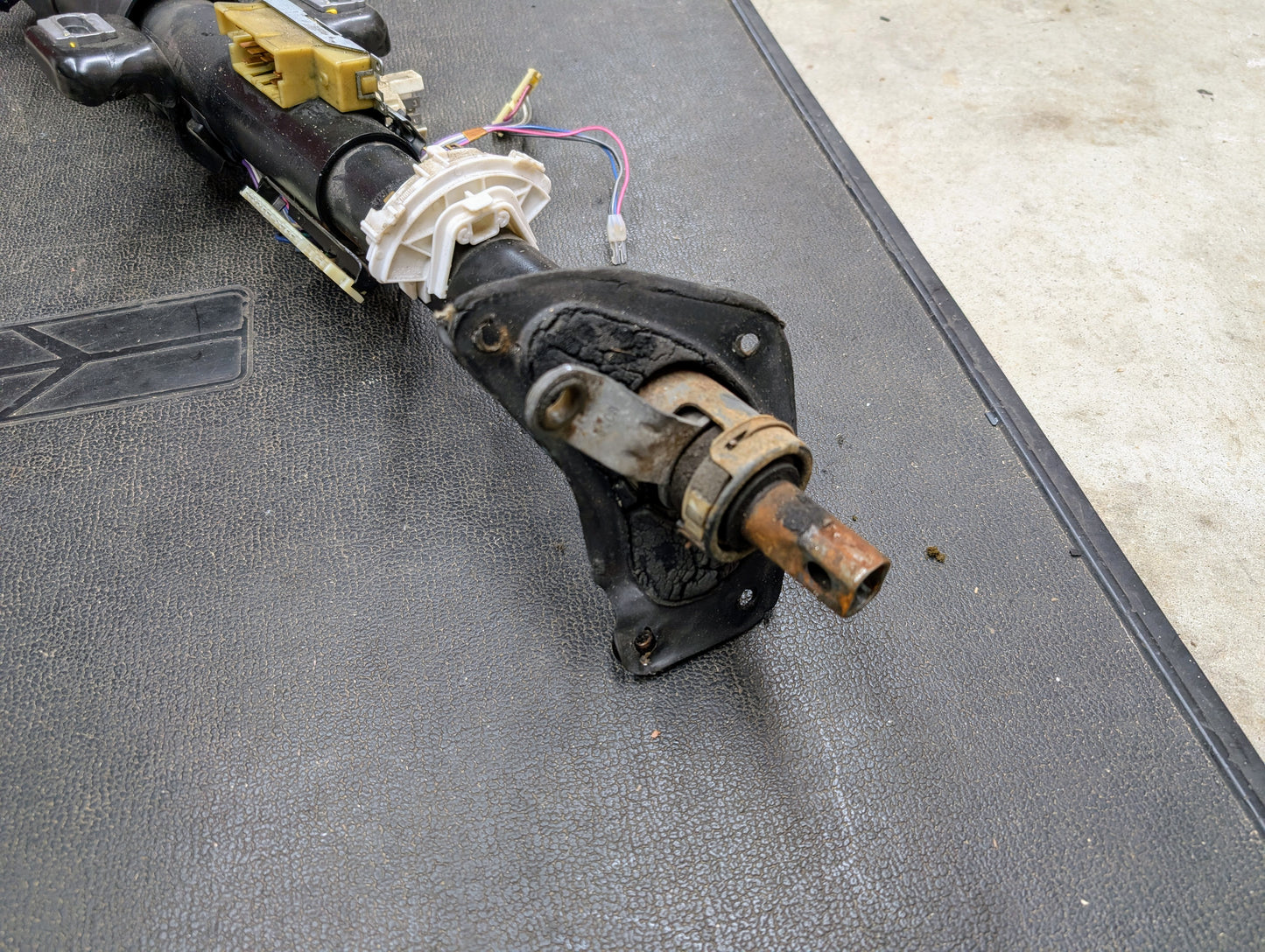 A close-up of a disassembled General Motors OEM Steering Column with Tilt and Cruise Control for 1988-1993 Chevy S10, Blazer, and more, showing exposed wires and metal parts on a textured black mat.