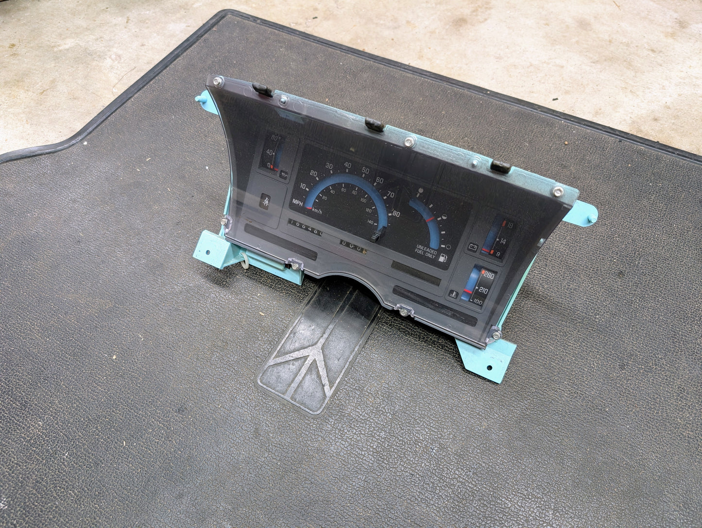 A General Motors OEM Instrument Cluster for 1986-1994 Chevy S10 Blazer sits on a textured black mat, showing a speedometer, fuel and temperature gauges, plus indicator lights. The background is concrete.