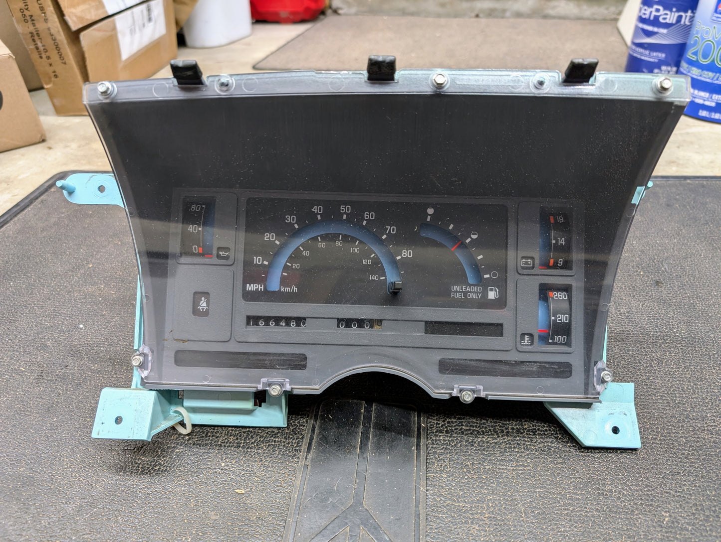 The General Motors OEM Instrument Cluster for 1986-1994 Chevy S10 Blazer features a speedometer, fuel gauge, and odometer under a clear cover with teal brackets, shown on a black mat in a garage.