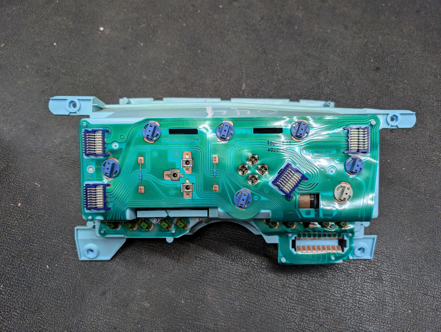 A close-up of a General Motors OEM Instrument Cluster for 1986-1994 Chevy S10 Blazer, featuring a green automotive circuit board with blue plastic connectors and electronic components on a black textured surface.