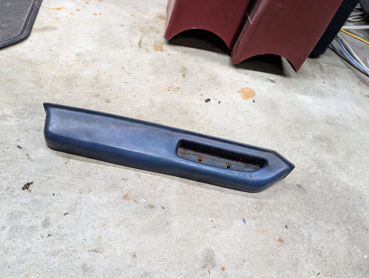 An OEM blue driver door panel armrest handle from General Motors for 1986–1993 Chevy S10 and Blazer rests on a concrete floor near maroon furniture and cables.