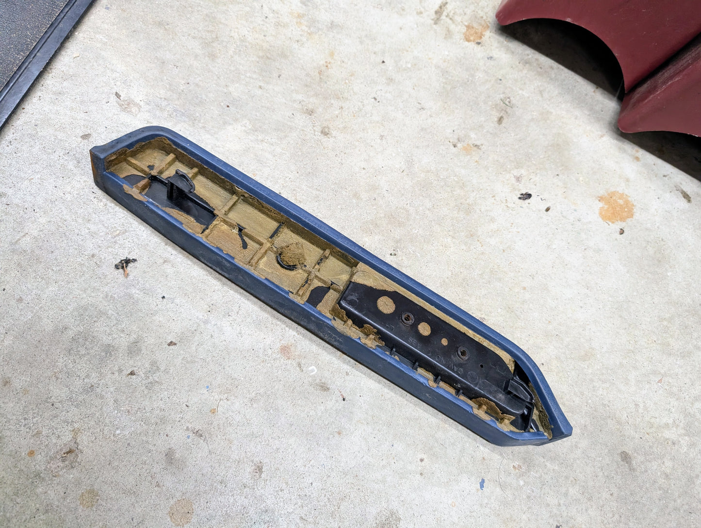 A used General Motors OEM Driver Door Panel Armrest Handle in blue for 1986-1993 Chevy S10 and Blazer, featuring rectangular shape, wear, damage, and a blue edge, is lying on a concrete floor.