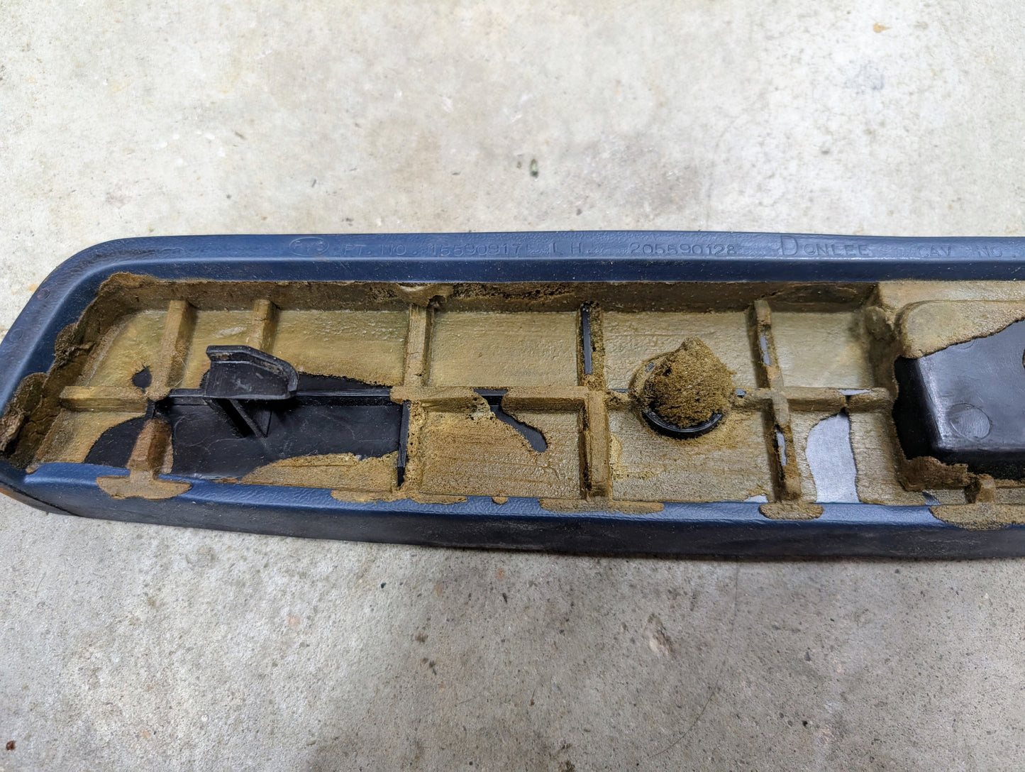 A close-up shows a blue OEM driver door panel armrest handle by General Motors for 1986-1993 Chevy S10, Blazer, and more, with accumulated dirt in its grooves and along the edges, resting on a concrete surface.