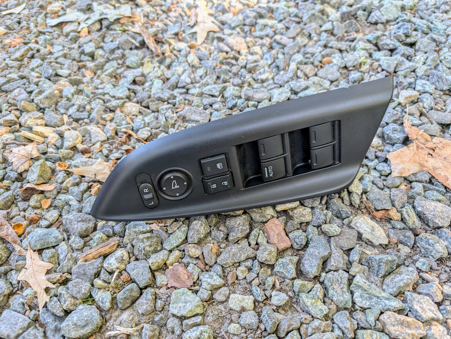 An OEM Driver Power Switch for 2015-2020 Honda Fit by Honda, featuring window and mirror adjustment buttons, lies on rocky ground with scattered dry leaves.