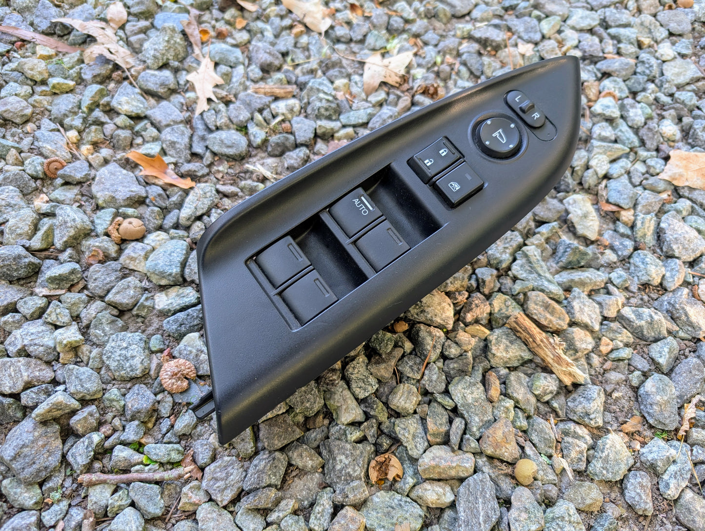 An OEM Driver Power Switch for 2015-2020 Honda Fit by Honda is lying on small gray gravel with dried leaves outdoors.