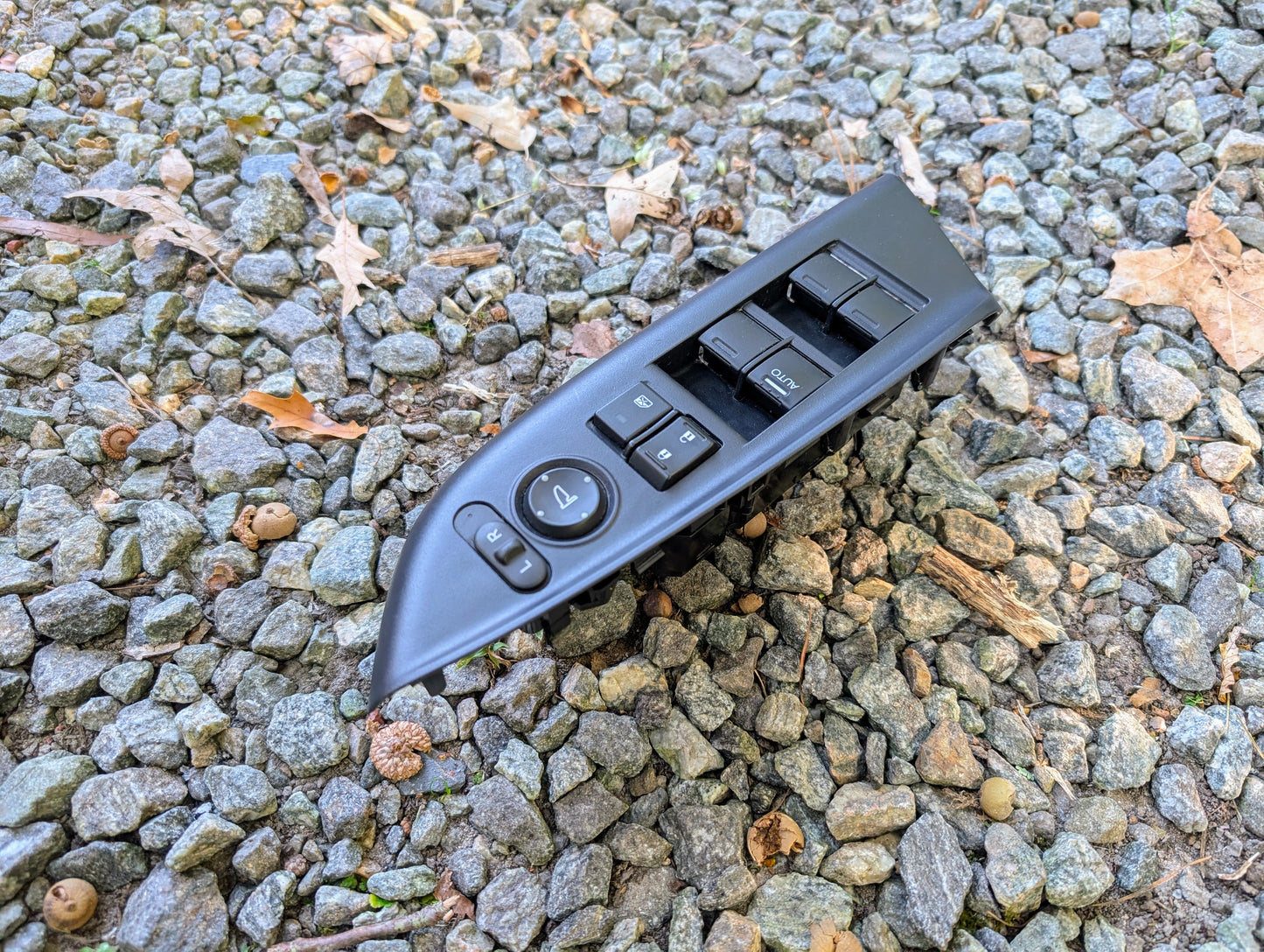 An OEM Driver Power Switch for 2015-2020 Honda Fit by Honda, featuring multiple buttons, is placed on a surface with small gray rocks and scattered brown leaves.