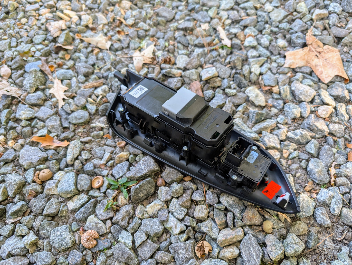 An OEM Driver Power Switch for 2015-2020 Honda Fit by Honda, featuring connectors and buttons, is placed on a gravel surface amid small stones and dry leaves.