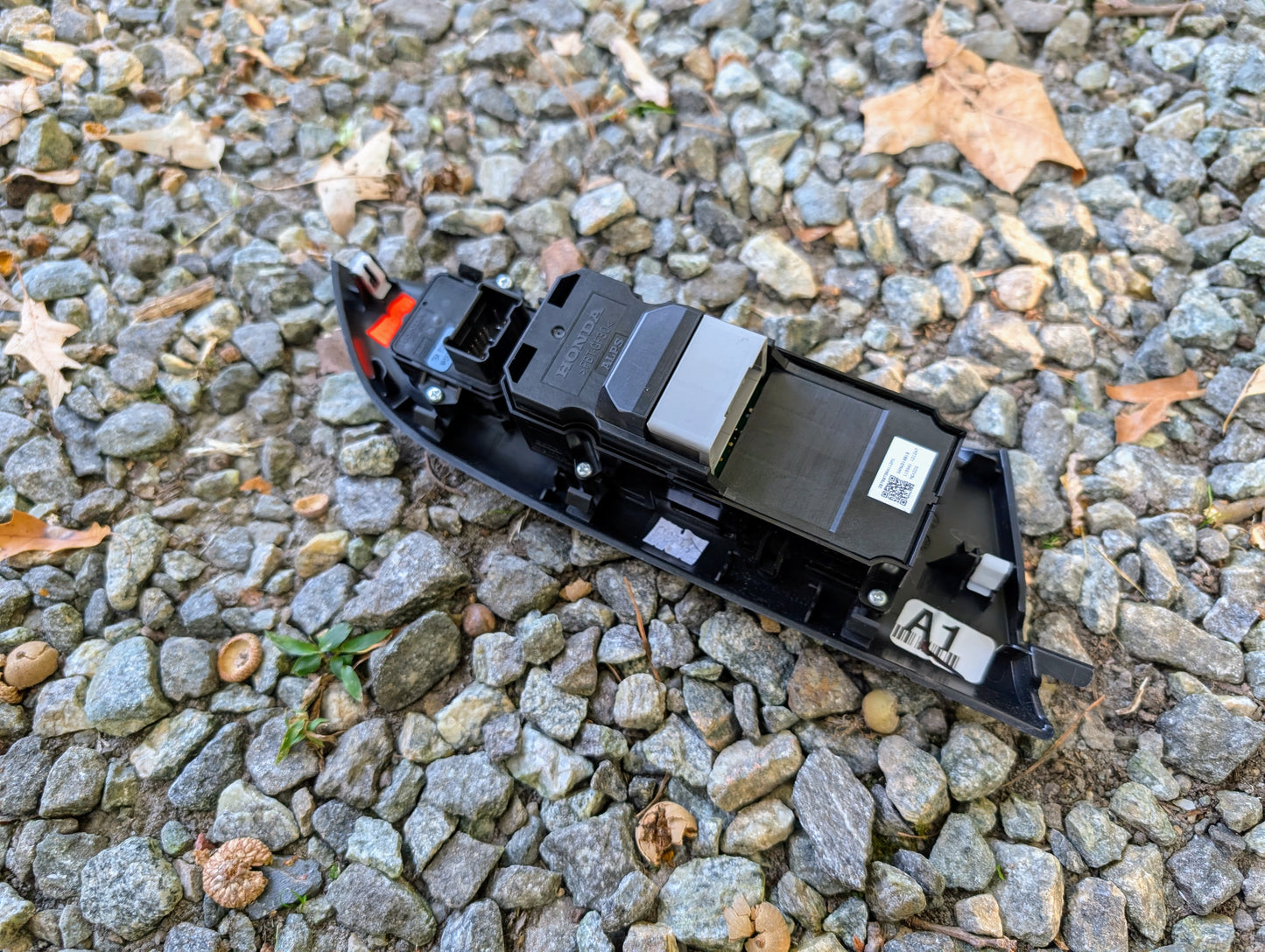 An OEM Driver Power Switch for 2015-2020 Honda Fit by Honda, featuring connectors and a barcode label, is pictured lying on gravel with scattered leaves and small pebbles.
