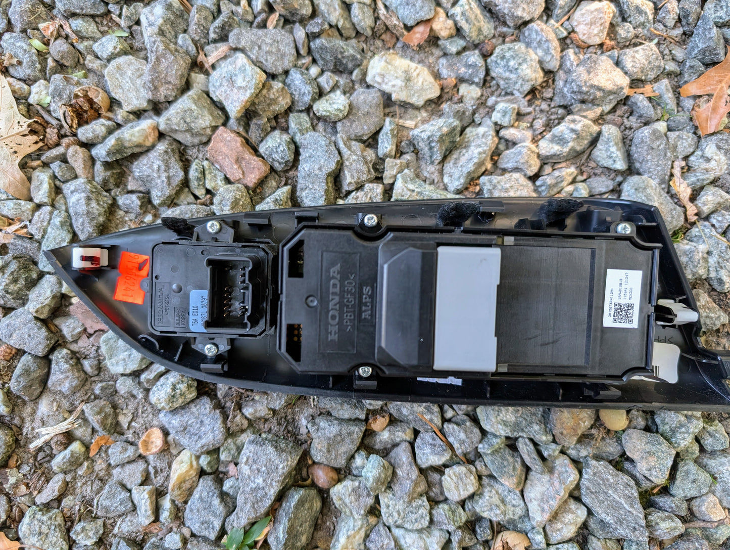 A Honda OEM Driver Power Switch for 2015-2020 Honda Fit rests on gray and brown rocks outdoors, its connectors and labels clearly visible.