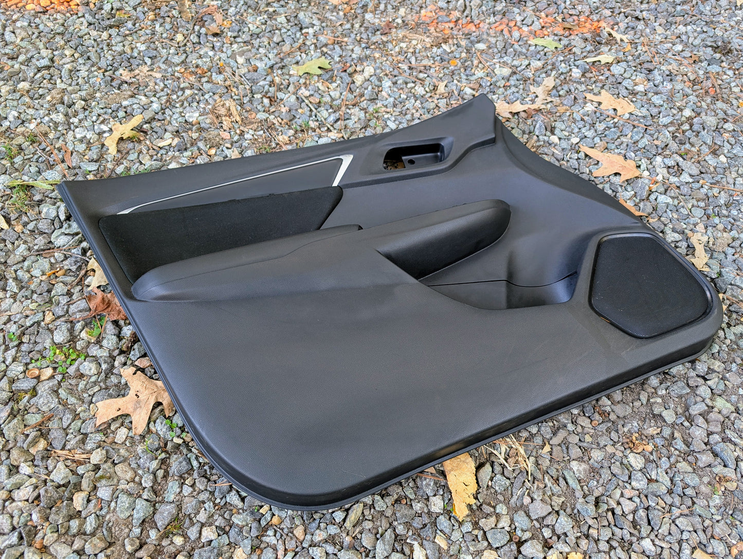 A Honda OEM Front Driver Power Door Panel for 2015-2020 Fit LX Trim Level rests on gravel outdoors, with brown leaves nearby. The black panel features armrest, handle cutouts, and speaker area.