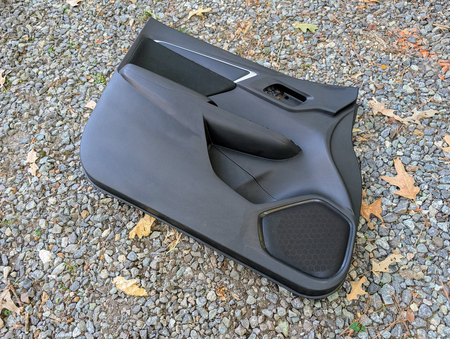 A Honda OEM Front Driver Power Door Panel for 2015-2020 Fit LX Trim Level rests on gravel, with scattered dry leaves around.