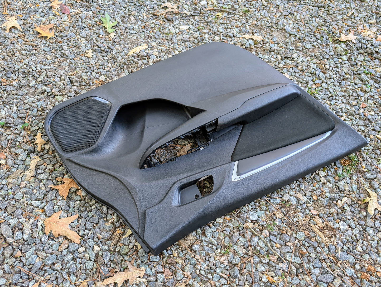 A Honda OEM Front Driver Power Door Panel for 2015-2020 Honda Fit LX Trim Level rests on a gravel surface, surrounded by scattered brown leaves.