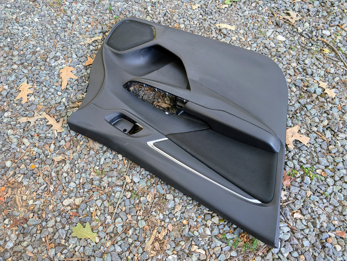 A Honda OEM Front Driver Power Door Panel for 2015-2020 Fit LX trim rests on gravel scattered with dry leaves, complete with handle and trim.