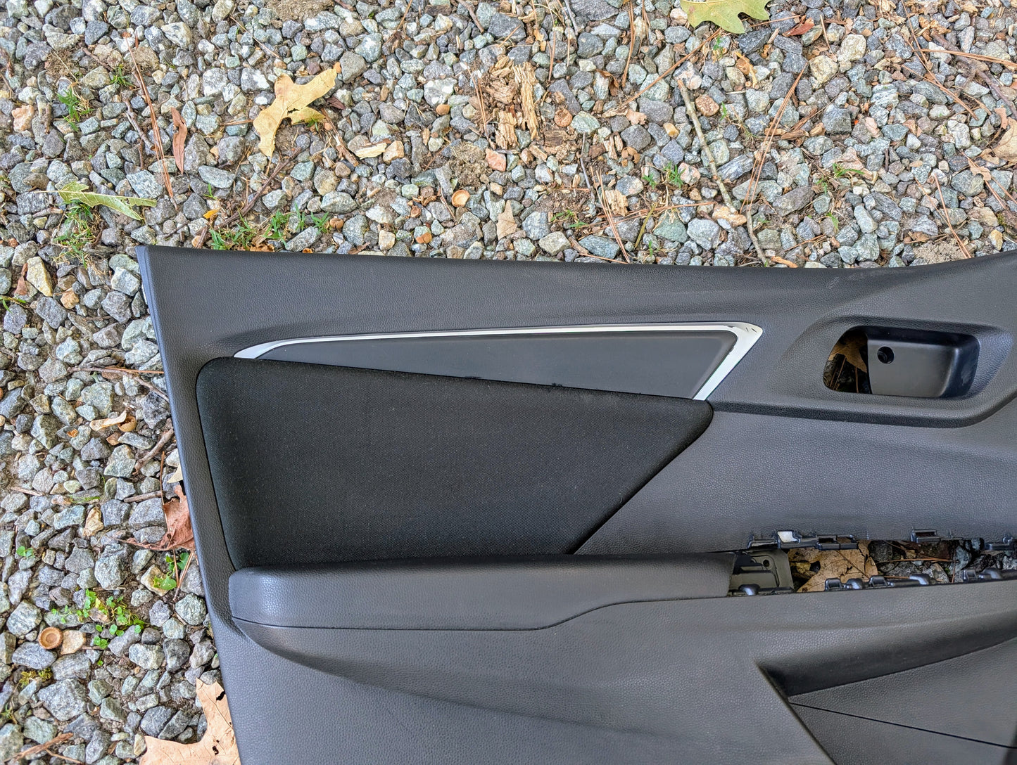 A Honda OEM Front Driver Power Door Panel for 2015-2020 Fit LX Trim Level, mostly black with a silver accent and partially disassembled, rests on gravel with scattered leaves.